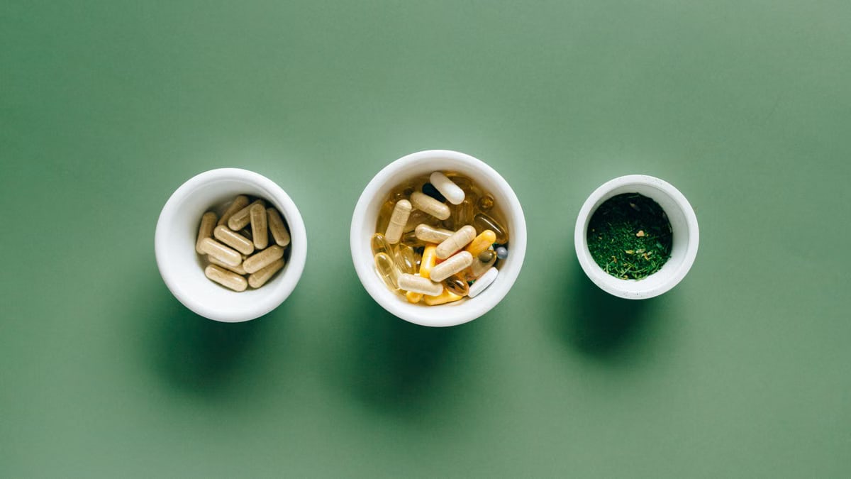 Top view of bowls with capsules, herbs, and supplements on green surface for healthcare concept.